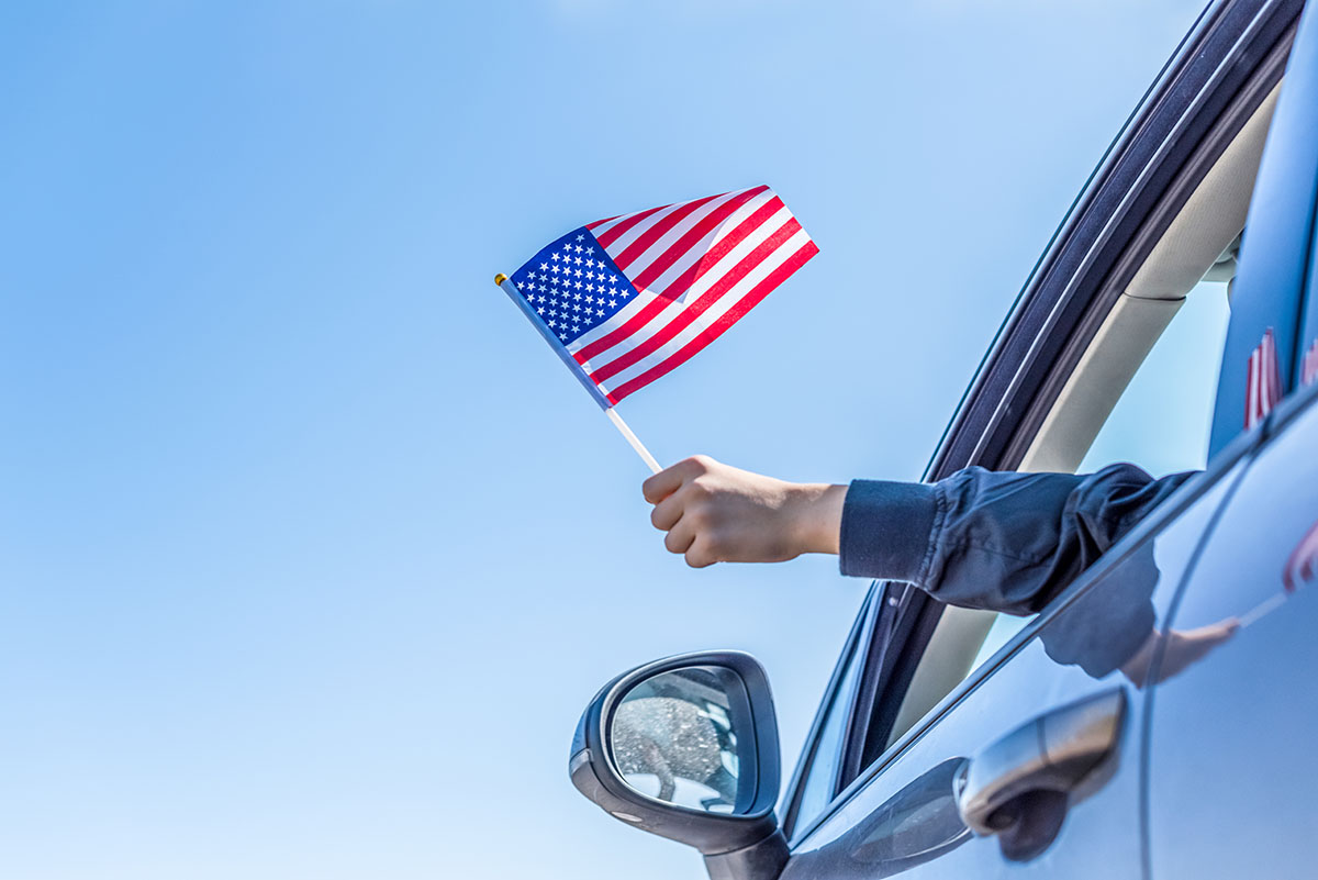 An arm waves an American flag through the window of a moving truck, with a blue sky in the background.