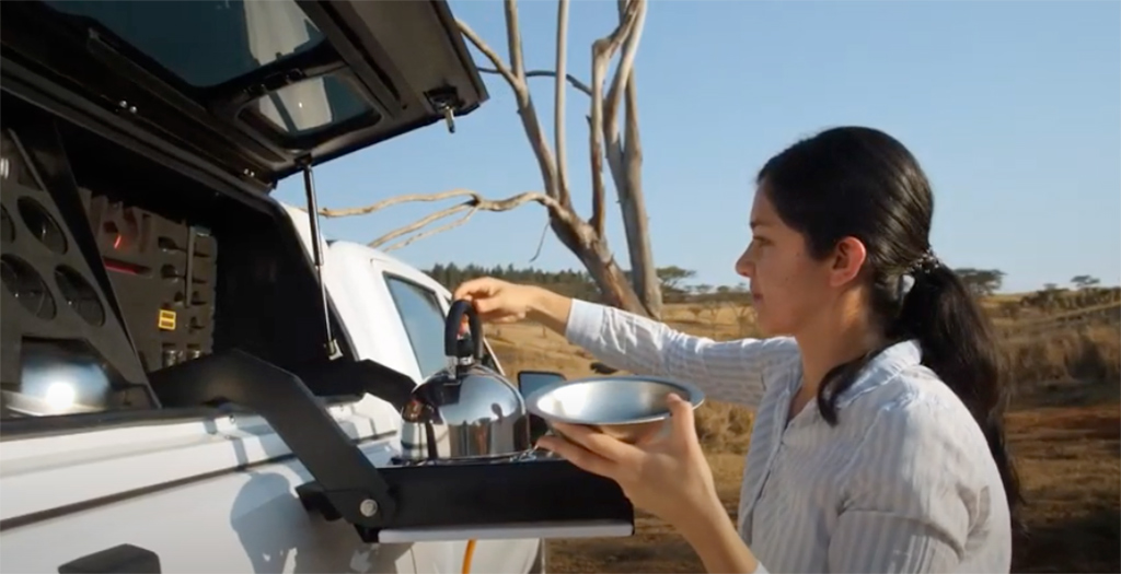 Woman reaching for tea kettle while at RSI SmartCap Gullwing Camp Kitchen mounted on truck cap on truck in wilderness.