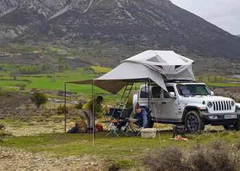 two people at a Mountainous campsite siting in camping chairs under the awning of a Jeep with a Thule Approach rooftop tent installed on top