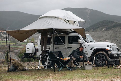 two people at a Mountainous campsite under the awning of a Jeep with a Thule Approach rooftop tent installed on top